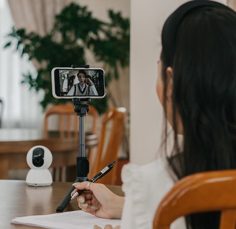 Woman participating in a virtual meeting via smartphone, taking notes in a cozy indoor setting.